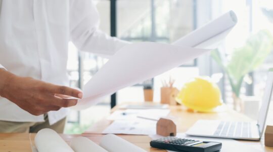 Construction professional reviews project plans with a calculator during early project cost planning.
