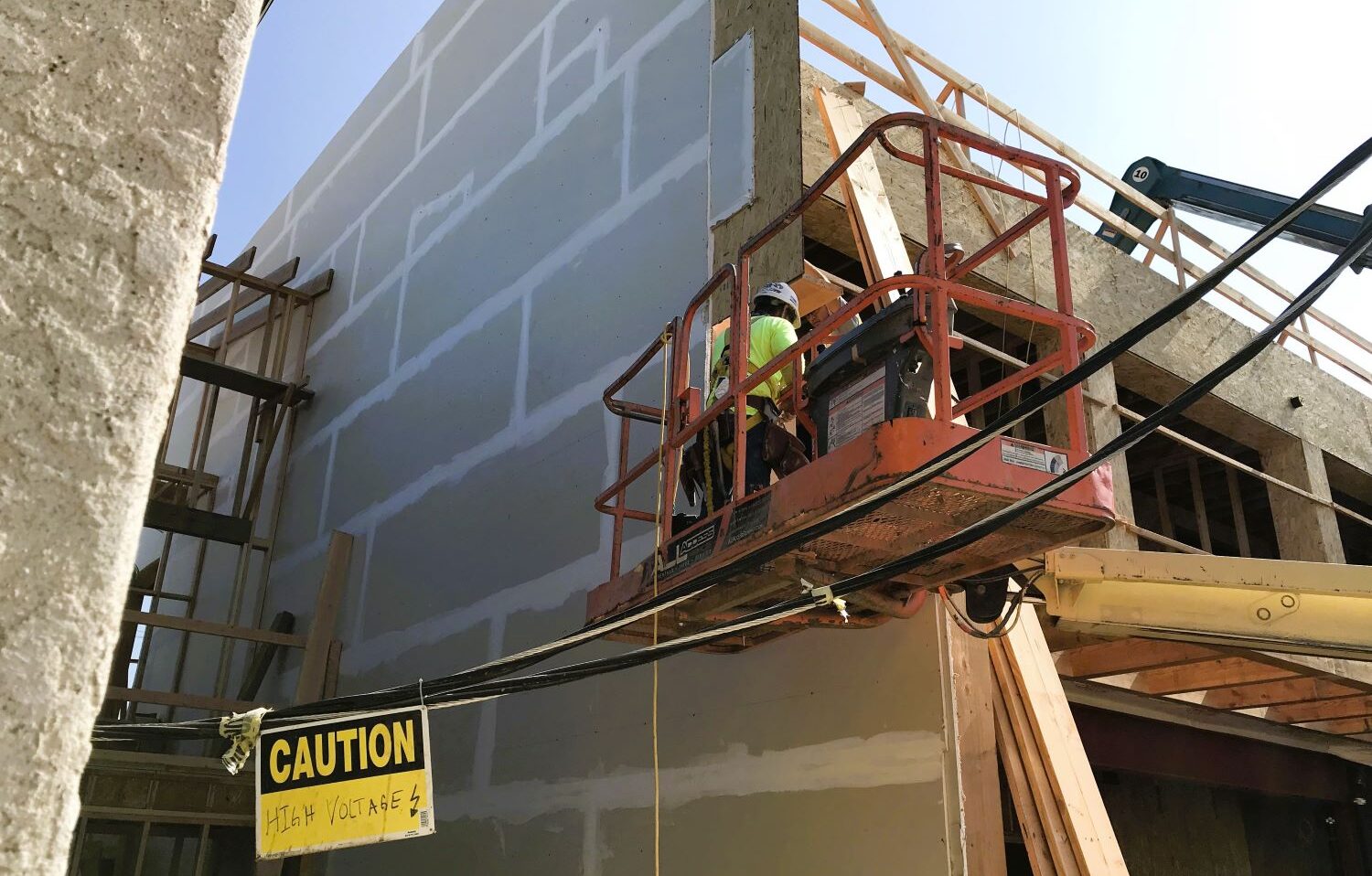 Construction worker on a lift installing exterior wall materials on a Level 3 Construction hotel project, with a high-voltage caution sign visible on site.