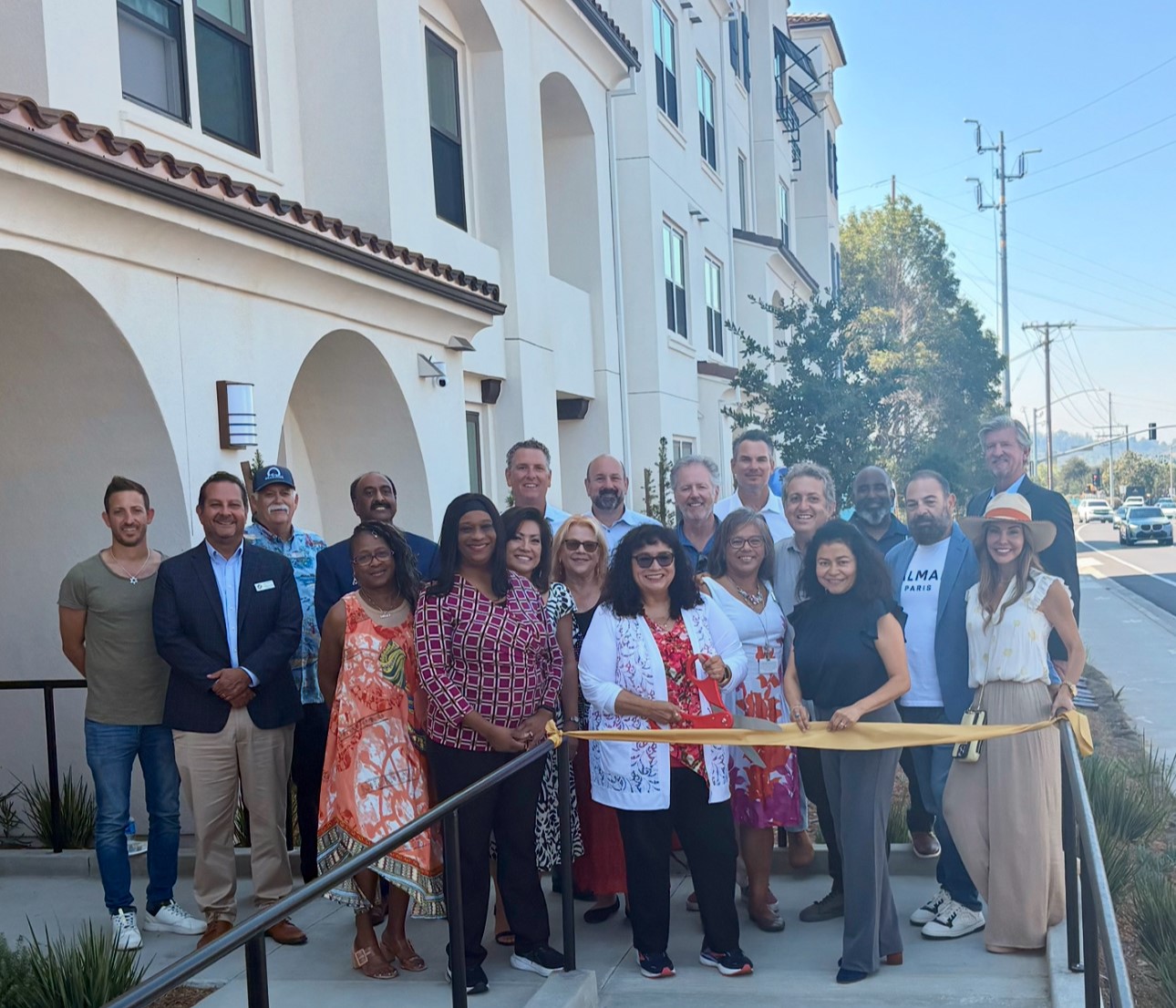 A group of about 50 people, including Oceanside city officials, developers, and community partners, gather in front of South River Village for the ribbon-cutting ceremony. The building's Spanish Mission-style exterior visible in the background.
