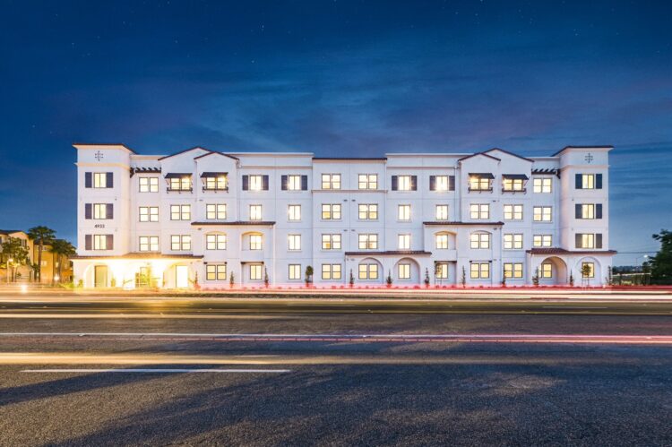 Exterior view of South River Village, a four-story Spanish Mission–style affordable housing community in Oceanside, CA, featuring stucco walls, a tile roof, and landscaped surroundings.