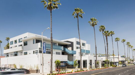 Exterior of The Flats, a mixed-use development with ground-floor retail and residential units above, along a palm-lined street in South Oceanside, California.