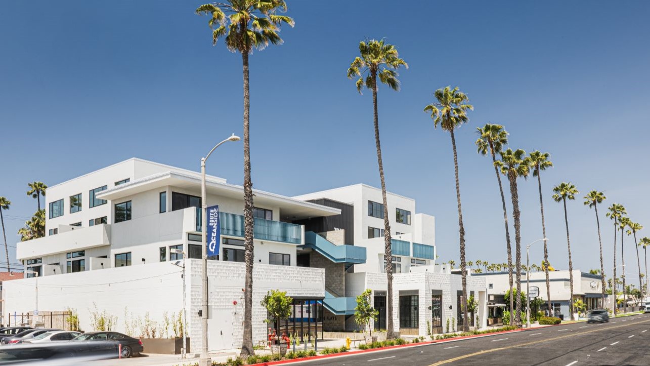 Exterior of The Flats, a mixed-use development with ground-floor retail and residential units above, along a palm-lined street in South Oceanside, California.
