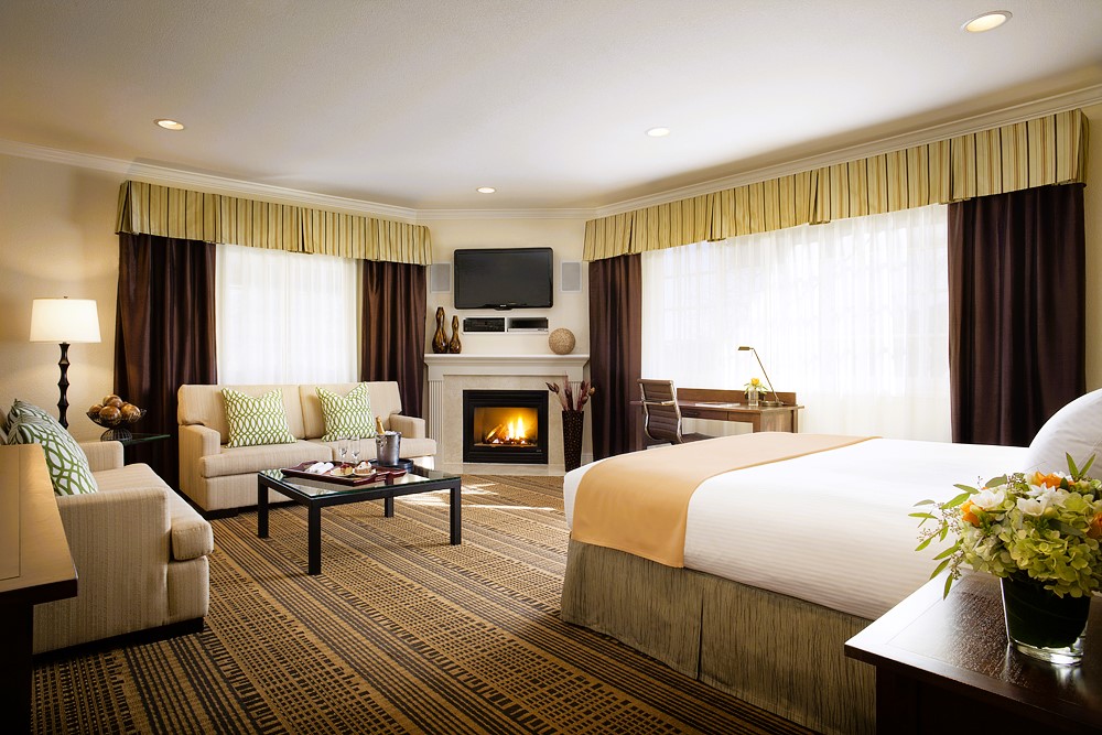 Spacious, modern hotel guest room with a king bed, seating area, desk, fireplace, and large windows with natural light.